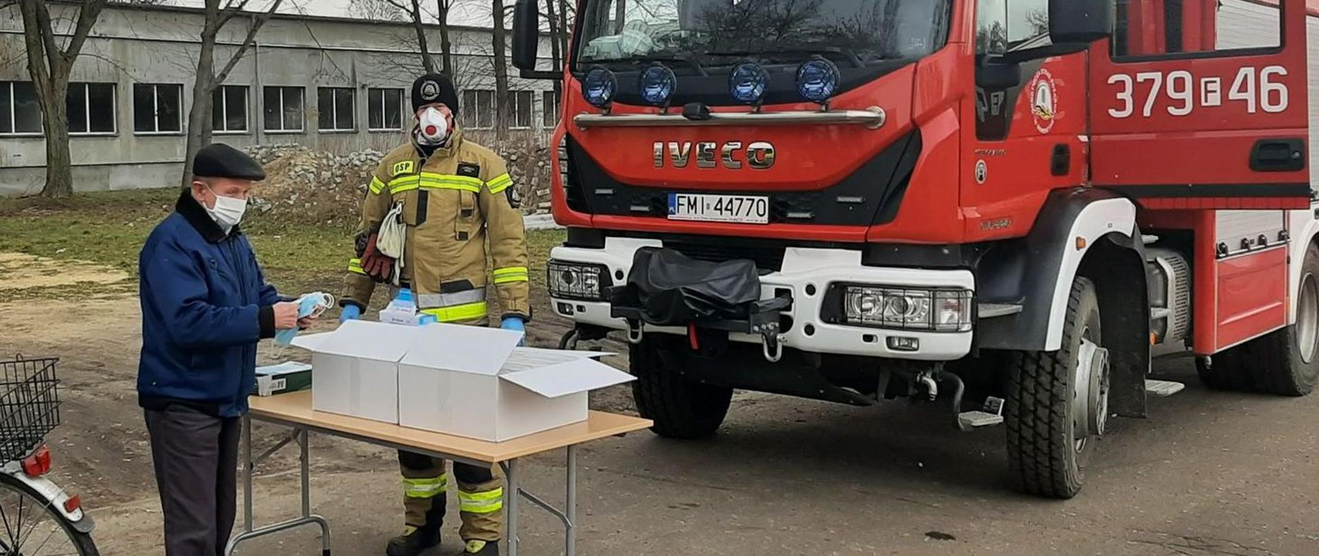 Zdjęcie przedstawia mieszkańca Skwierzyny odbierającego maseczki od strażaka OSP na punkcie wydawczym zorganizowanym w Skwierzynie. Obok stoi strażak, za nim pojazd pożarniczy.