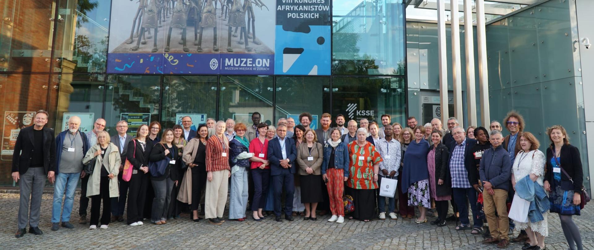 Zdjęcie grupowe 48 uczestników VIII Kongresu Afrykanistów w Żorach; niektórzy z nich noszą przepustki; prawie wszyscy uśmiechaja się do zdjęcia; w tle budynek Muzeum w Żorach; nowoczesna szklana fasada; budynek jest dwupiętrowy; na oknach wiszą plakaty; największy plakat informuje, że Muzeum w Żorach obchodzi 25 lat; mniejszy plakat informuje o VIII kongresie Afrykanistów w Żorach; mniejsze plakaty wywieszone na poziomie parteru informują o organizowanych wydarzeniach; teren przed Muzeum wybrukowany jest szarą kostką; ciemniejszą szarą kostką wybrukowane są elementy dekoracyjne