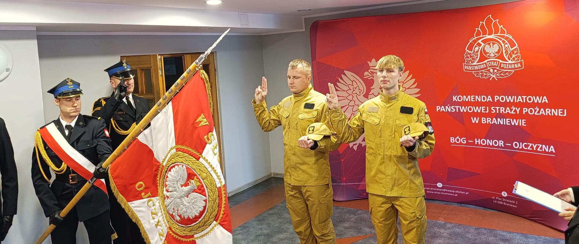 Na zdjęciu uwieczniono uroczystość ślubowania funkcjonariuszy Komendy Powiatowej Państwowej Straży Pożarnej w Braniewie. Dwóch strażaków, ubranych w umundurowanie specjalne, składa rotę ślubowania w obecności pocztu sztandarowego oraz kadry komendy. Uroczystość odbyła się w siedzibie KP PSP w Braniewie i miała oficjalny, podniosły charakter.