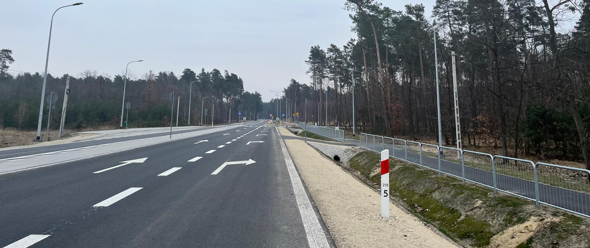 Fotografia przedstawia prostą, asfaltową drogę z wyraźnie oznaczonymi pasami ruchu i strzałkami kierunkowymi. Po lewej stronie drogi znajdują się latarnie uliczne, a po prawej stronie widoczny jest chodnik oddzielony metalowym ogrodzeniem. W tle widać las z drzewami iglastymi i liściastymi. Na pierwszym planie, po prawej stronie, znajduje się biały słupek z czerwonym paskiem i numerem "5". Droga jest pusta, bez ruchu pojazdów, co może sugerować, że jest to nowo wybudowana lub mało uczęszczana trasa.