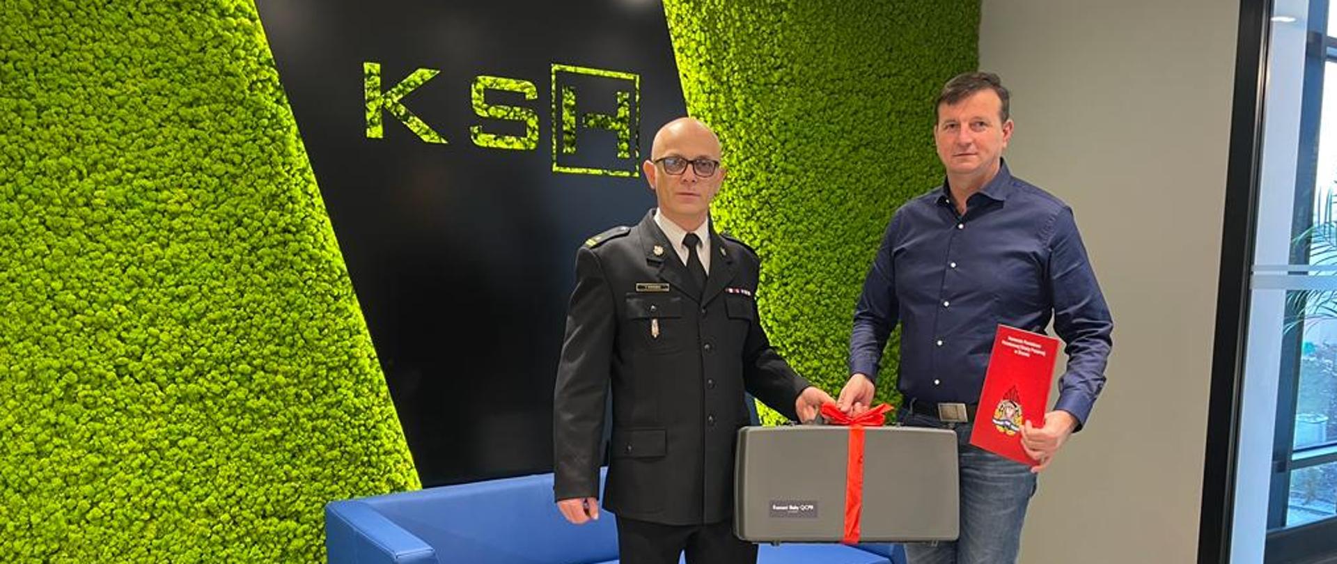na zdjęciu komendant powiatowy PSP wraz z prezesem firmy KSH podczas przekazania fantom którego trzymają na tle loga firmy KSH na zielonym tle 