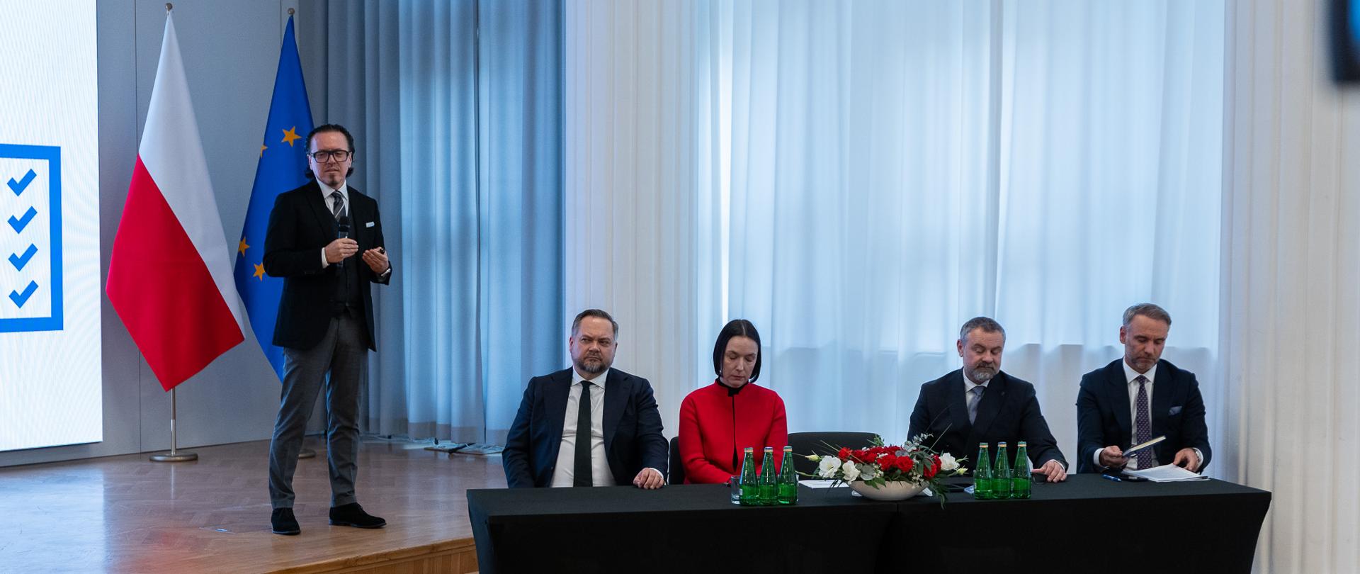 Minister Wojciech Balczun stoi na tle flag, trzyma w dłoni mikrofon. Wiceministrowie i Dyrektor Generalny siedzą za stołem.