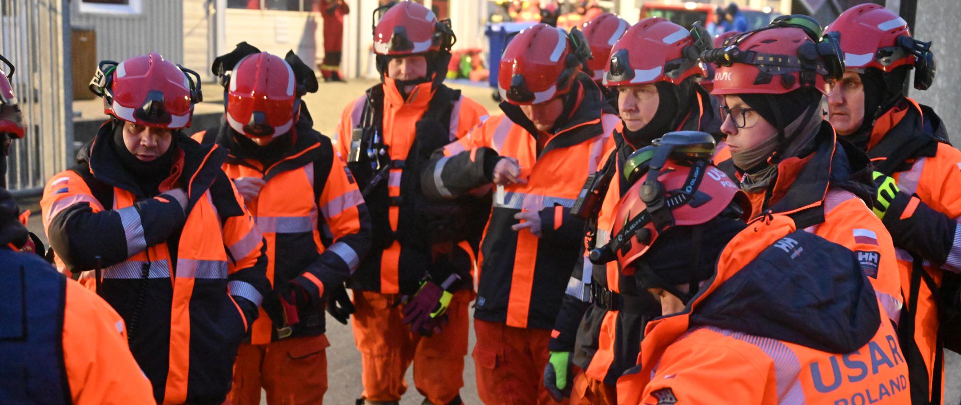 Grupa ratowników USAR Poland w kaskach i pomarańczowych kurtkach uczestniczy w odprawie przed rozpoczęciem działań.