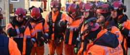 Grupa ratowników USAR Poland w kaskach i pomarańczowych kurtkach uczestniczy w odprawie przed rozpoczęciem działań.