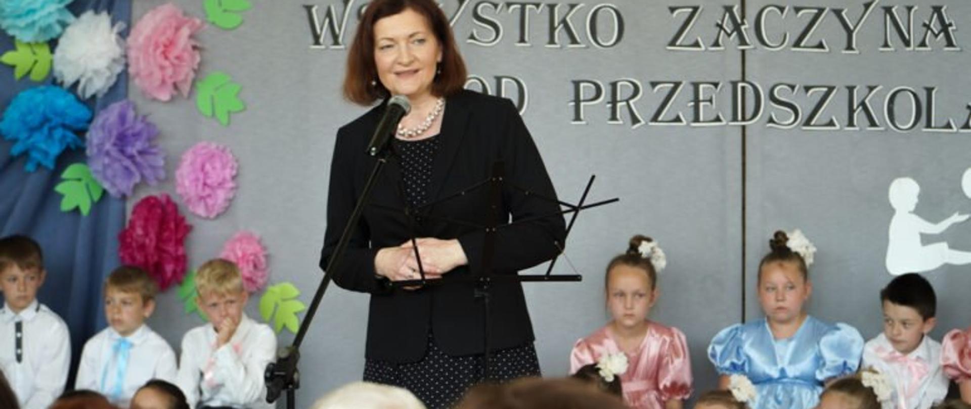 Wojewoda podkarpacki Ewa Leniart przemawia podczas otwarcia budynku przedszkola przy Szkole Podstawowej w Futomie. W tle dzieci przedszkolne. 