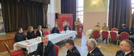 Strażacy ubrani w granatowe mundury galowe, oraz pisakowe mundury siedzą przy stolikach, w tyle rol-ap z logiem Państwowej Straży Pożarnej na tylnej ścianie kotary oraz stół prezydialny. Przy mównicy stoi Komendant Miejski w piaskowym mundurze, przed sobą ma mikrofon