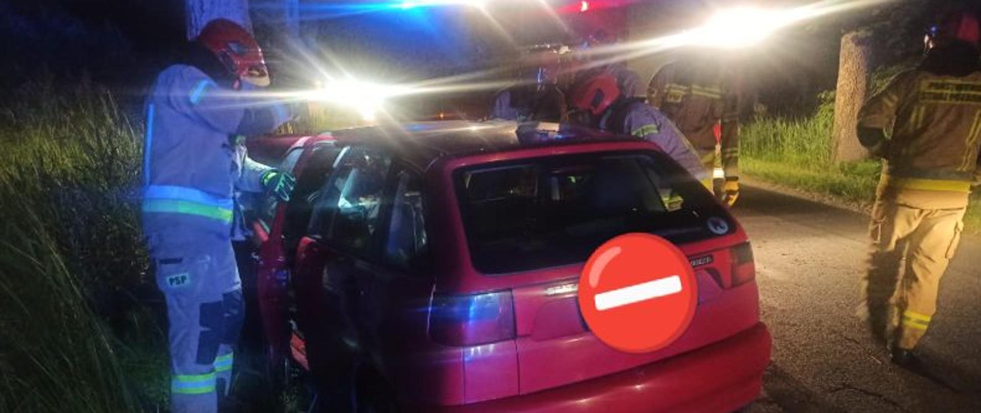Zdjęcie wykonane w nocy. Oświetlone błyskami samochodów gaśniczych. Auto stoi uderzone przodem w drzewo. Auto koloru czerwonego. Przy aucie strażacy w piaskowych mundurach.