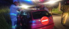 Zdjęcie wykonane w nocy. Oświetlone błyskami samochodów gaśniczych. Auto stoi uderzone przodem w drzewo. Auto koloru czerwonego. Przy aucie strażacy w piaskowych mundurach.