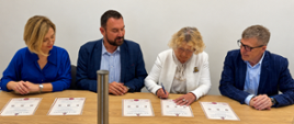 Cztery osoby uczestniczą w oficjalnym spotkaniu przy drewnianym stole w sali konferencyjnej. Jedna z uczestniczek podpisuje dokument, pozostali obserwują przebieg podpisania. Na stole leżą przygotowane akty oraz długopis.