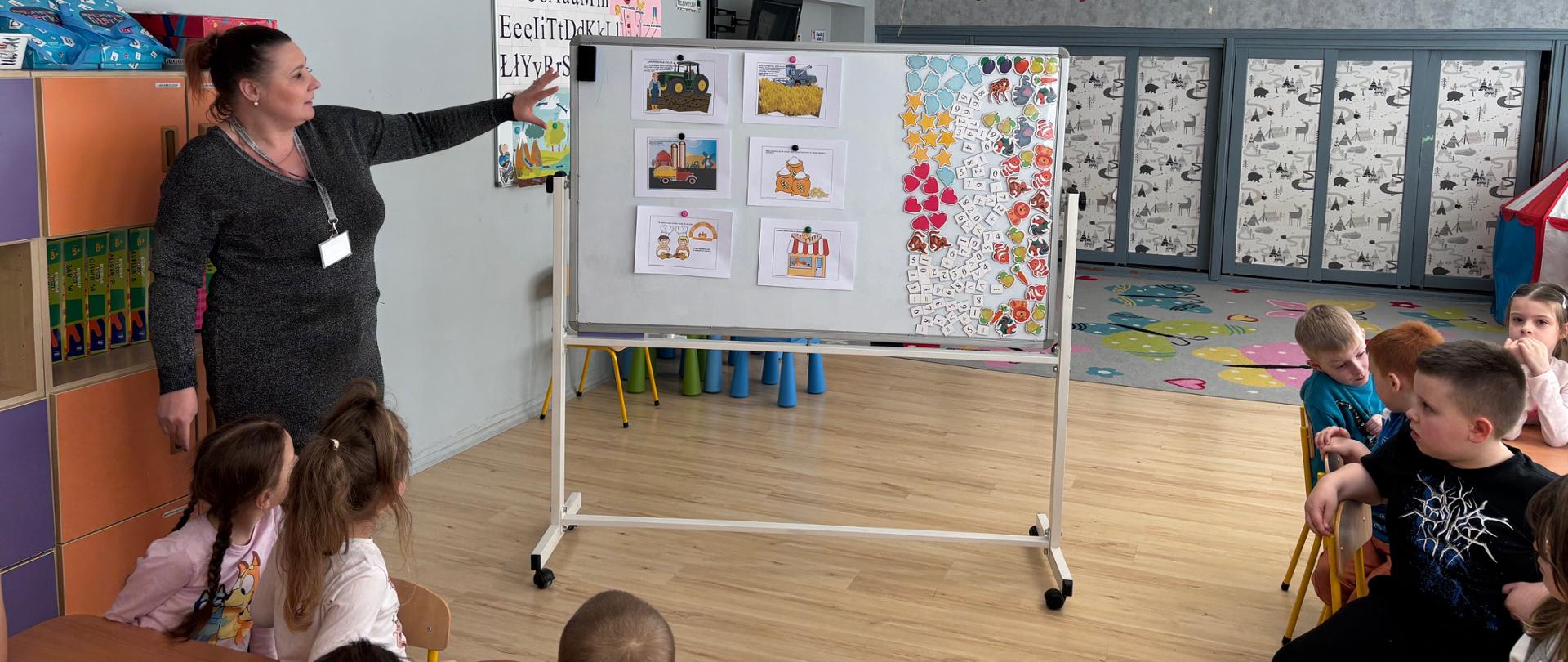 Inspektor WIORiN stojący przy tablicy na sali wraz z dziećmi siedzącymi przy stolikach.