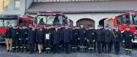 Wspólne zdjęcie strażaków w tle samochody pożarnicze oraz budynek komendy.
