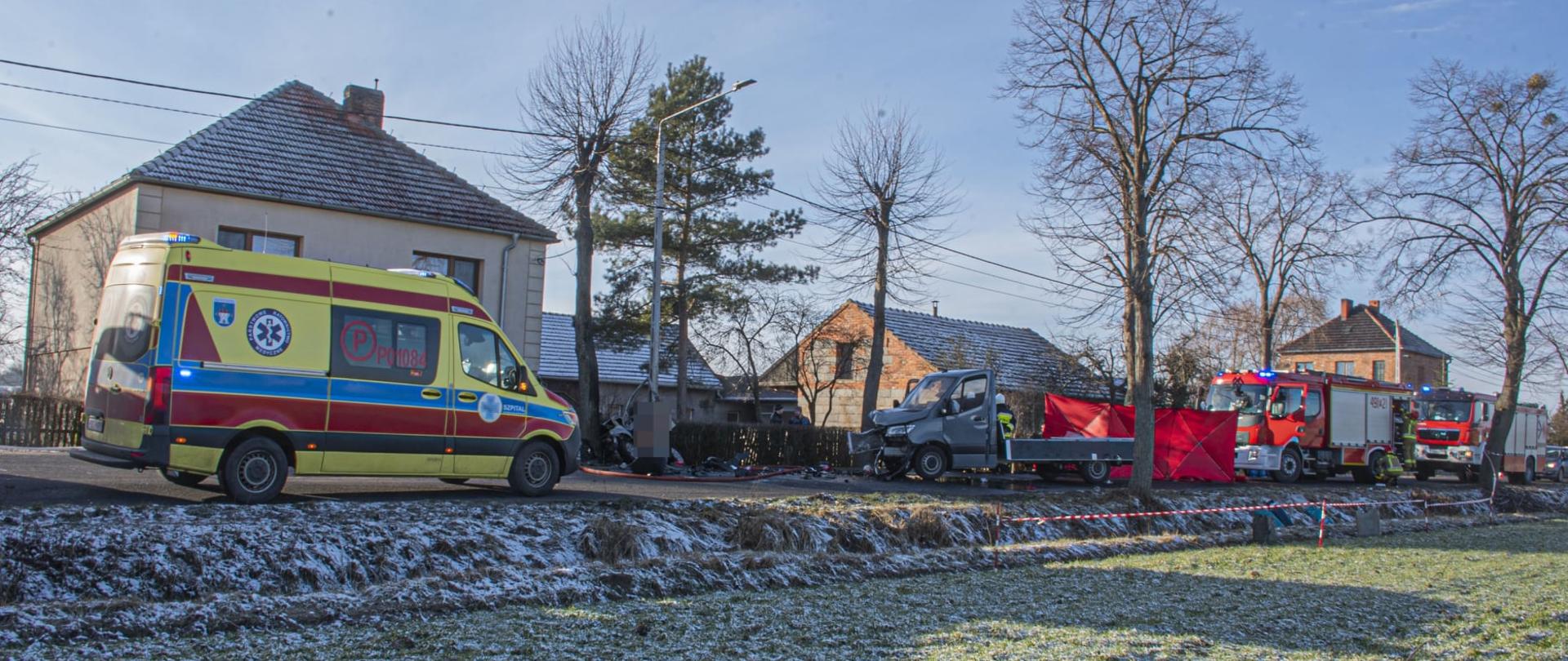 Zdjęcie przedstawia karetkę pogotowia ratunkowego koloru żółtego, rozbity samochód dostawczy oraz samochody straży pożarnej koloru czerwonego. Miejsce prowadzonych działań zostało odgrodzone taśmą ostrzegawczą oraz parawanem ratowniczym koloru czerwonego.