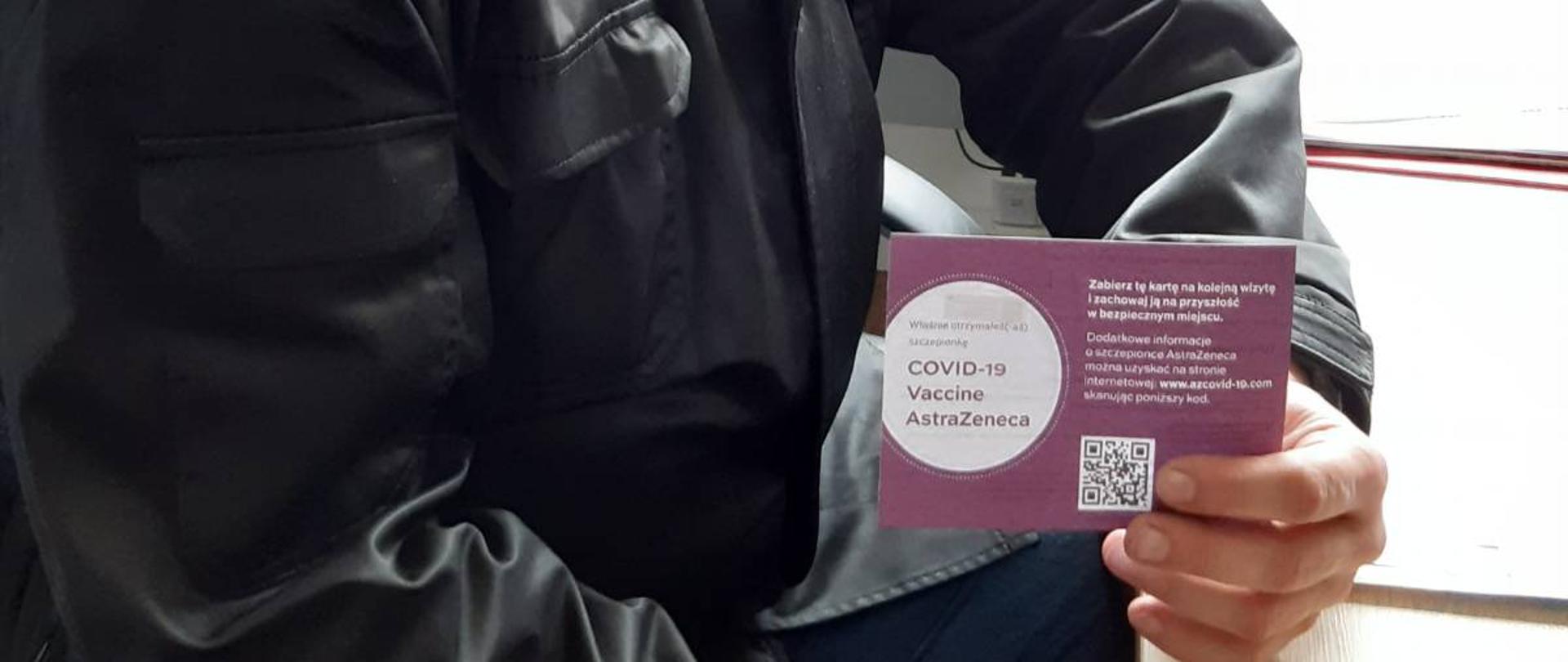 Zdjęcie przedstawia strażaka w czarnym mundurze sztabowym siedzącego przy biurku w pomieszczeniu. W ręku trzyma bordowo-białą kartkę z napisem „COVID-19 Vaccine AstraZeneca”. W tle okno, telefony, kwiat i radiostacja. 