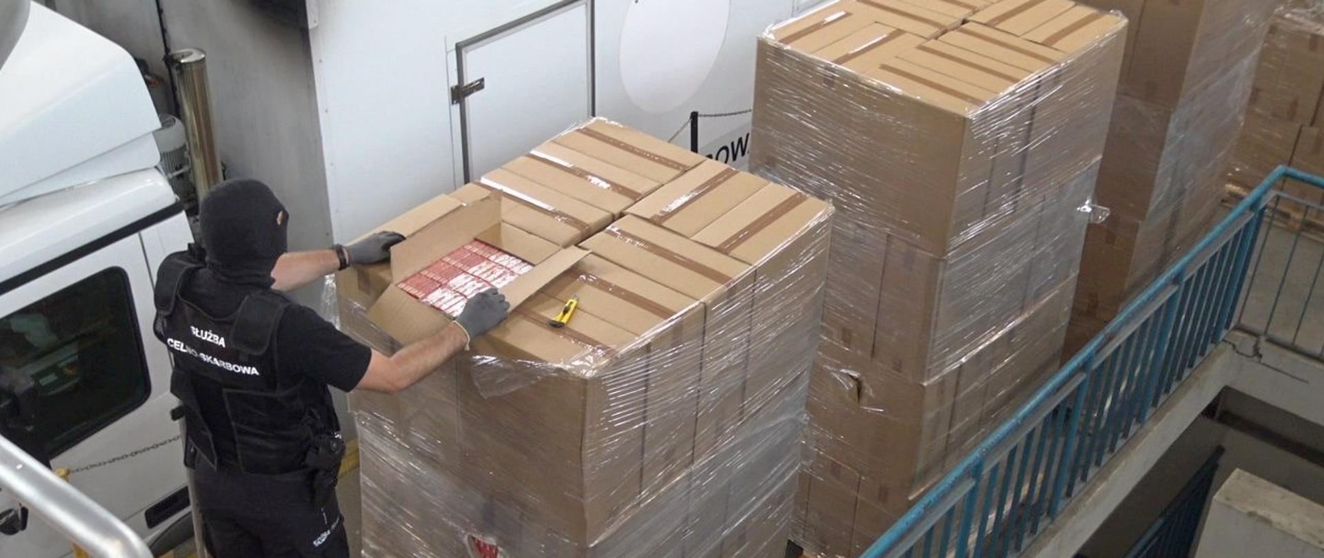 Hala, palety z kartonami z papierosami, funkcjonariusz KAS przy jednym z otwartych kartonów.