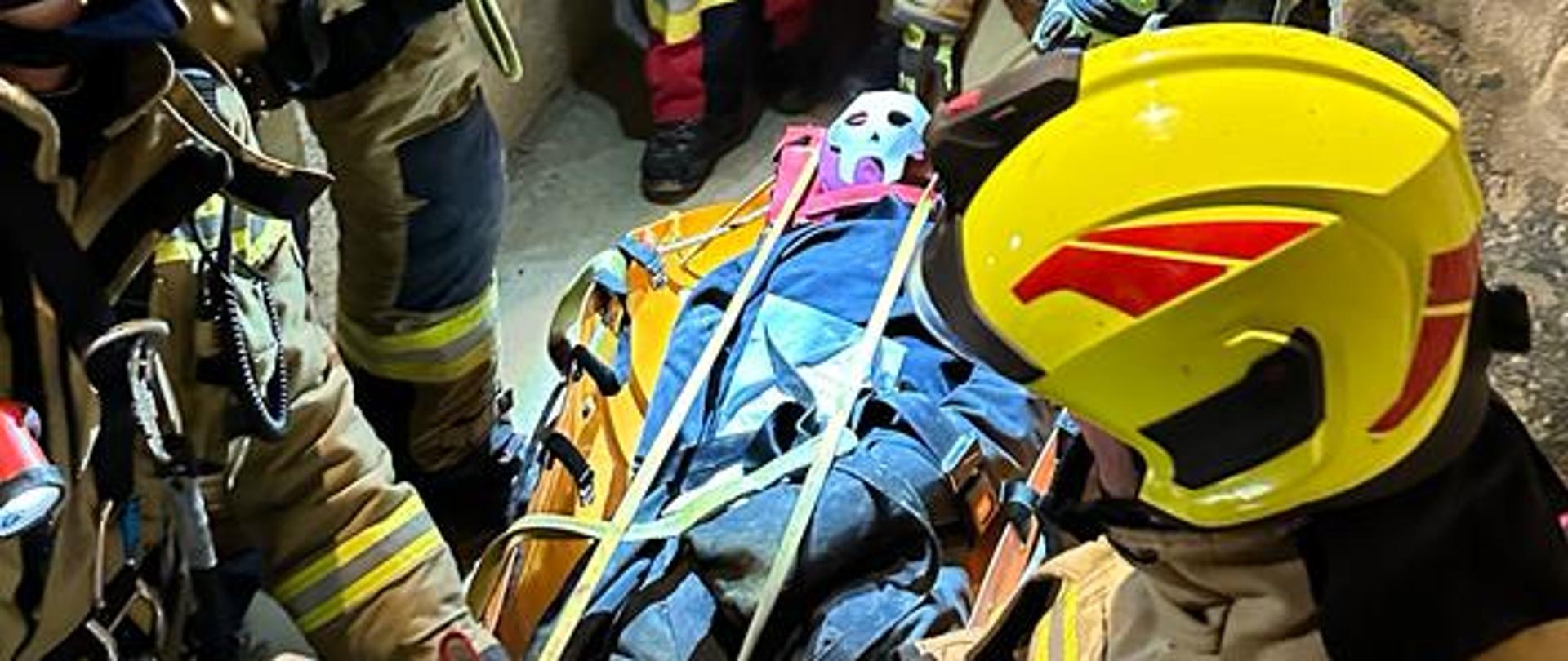 Strażacy podczas ćwiczeń w wąskim przejściu kopalni. Na podłodze widoczna jest osoba poszkodowana na noszach ratowniczych przygotowana do ewakuacji.