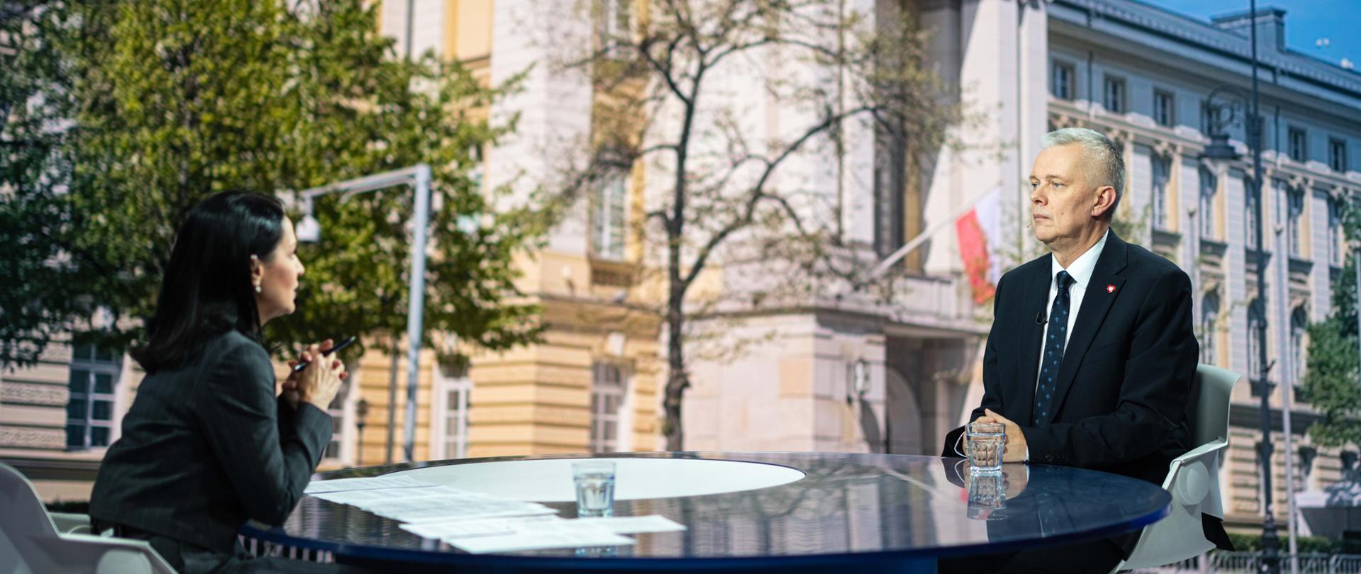 Minister Tomasz Siemoniak po prawej stronie kadru i dziennikarka Diana Rudnik po lewej podczas rozmowy przy okrągłym stole w studiu telewizyjnym. Na stole leżą dokumenty i stoją szklanki z wodą, a w tle widoczne jest zdjęcie KPRM.