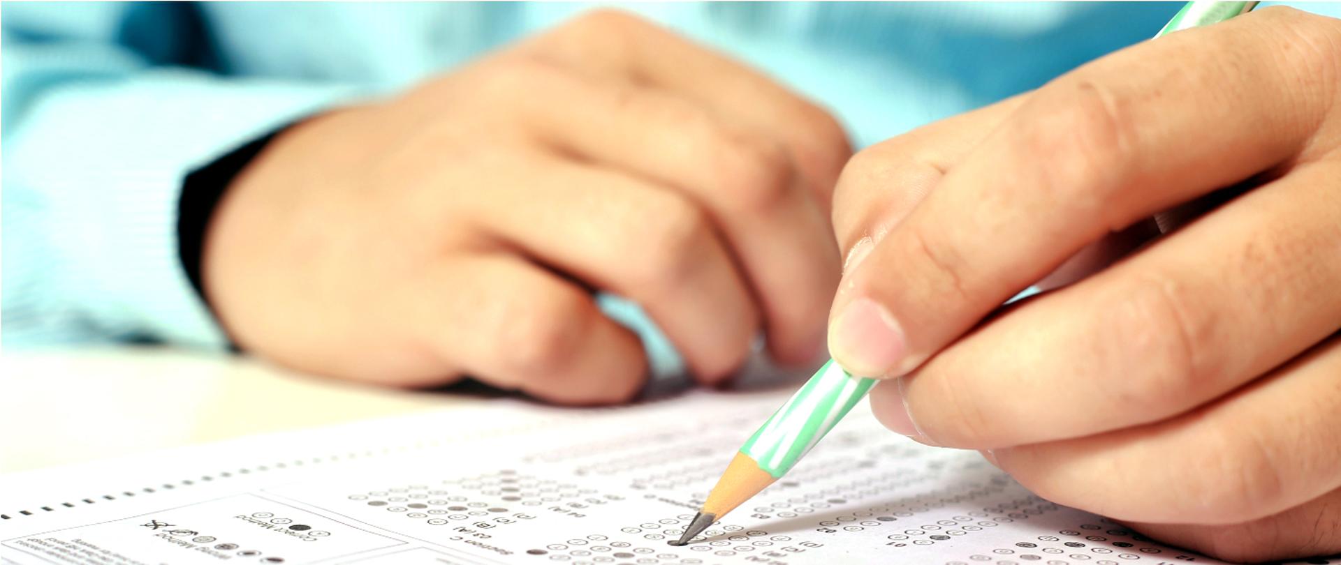 Grafika przedstawia ręce ucznia wypełniającego ołówkiem akrusz egzaminacyjny