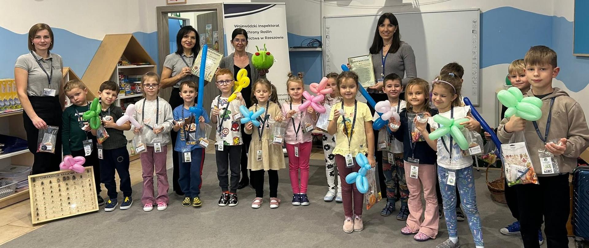 Grupa dzieci stoi w Sali. Ściany są niebiesko-białe. Dzieci ubrane sa na kolorowo i w dłoni trzymają woreczki z cukierkami i gadżetami. Na ścianach są kolorowe obrazki.Za dziećmi stoją cztery kobiety. Wszyscy się uśmiechają.