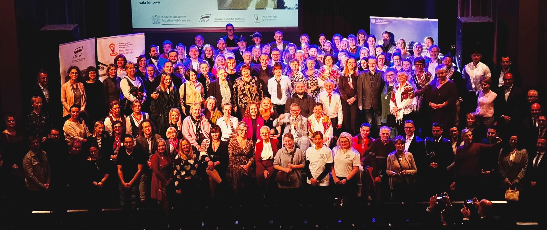 Szeroki kadr, zdjęcie grupowe. Ujęcie z góry. Uczestnicy konferencji regionalnej pozują do wspólnego zdjęcia stojąc na scenie sali kinowej. Przed uczestnikami widoczne rzędy pustych foteli. W tle widoczny ekran, na którym wyświetlona jest grafika informująca o wydarzeniu, z symboliczną ilustracją. Ilustracja przedstawia grupę ludzi podających sobie uniesione w górę dłonie. Obok widoczny jest tytuł: Konferencja regionalne Moc Małych Społeczności, Żory, województwo śląskie, oraz szczegóły spotkania: 12 listopada, godzina 14, Miejski Ośrodek Kultury w Żorach, sala kinowa. Na grafice widoczne są także loga: Komitetu do spraw Pożytku Publicznego, Narodowego Instytutu Wolności oraz programu Moc Małych Społeczności. Za uczestnikami konferencji widoczne są również fragmenty banerów promocyjnych.