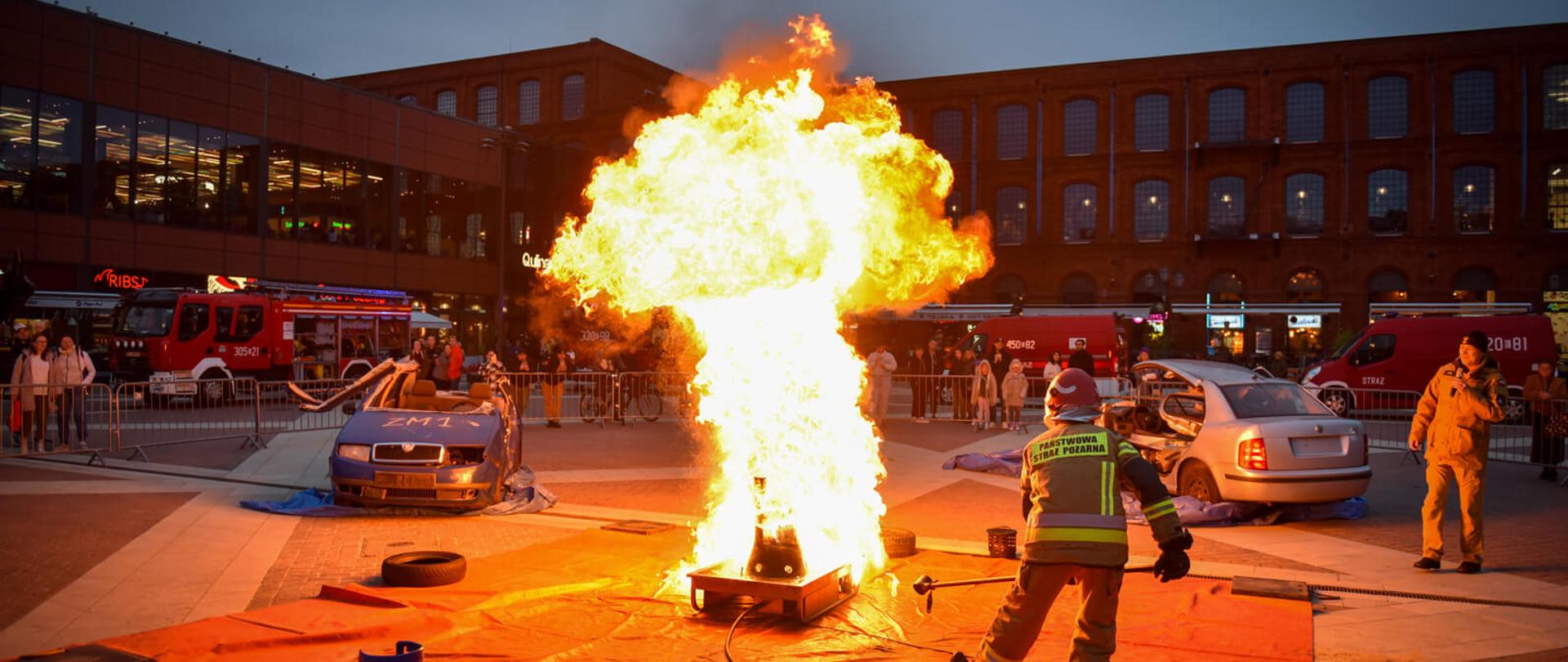 Nocny pokaz z ognia na placu. Ogromna, jasna kula ognia unosi się w powietrzu. Strażak w odblaskowym kombinezonie stoi blisko ognia, patrząc w jego kierunku. W tle widać dwa samochody, wóz strażacki oraz budynki.