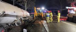 W rowie przewrócona biała cysterna na drodze asfaltowej strażacy w ubraniach specjalnych z tył samochody pożarnicze oraz żółta koparka.