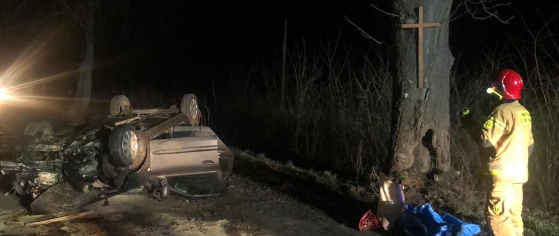 Samochód osobowy leżący na dachu, obok strażak stojący przy drzewie, na którym wisi krzyż.