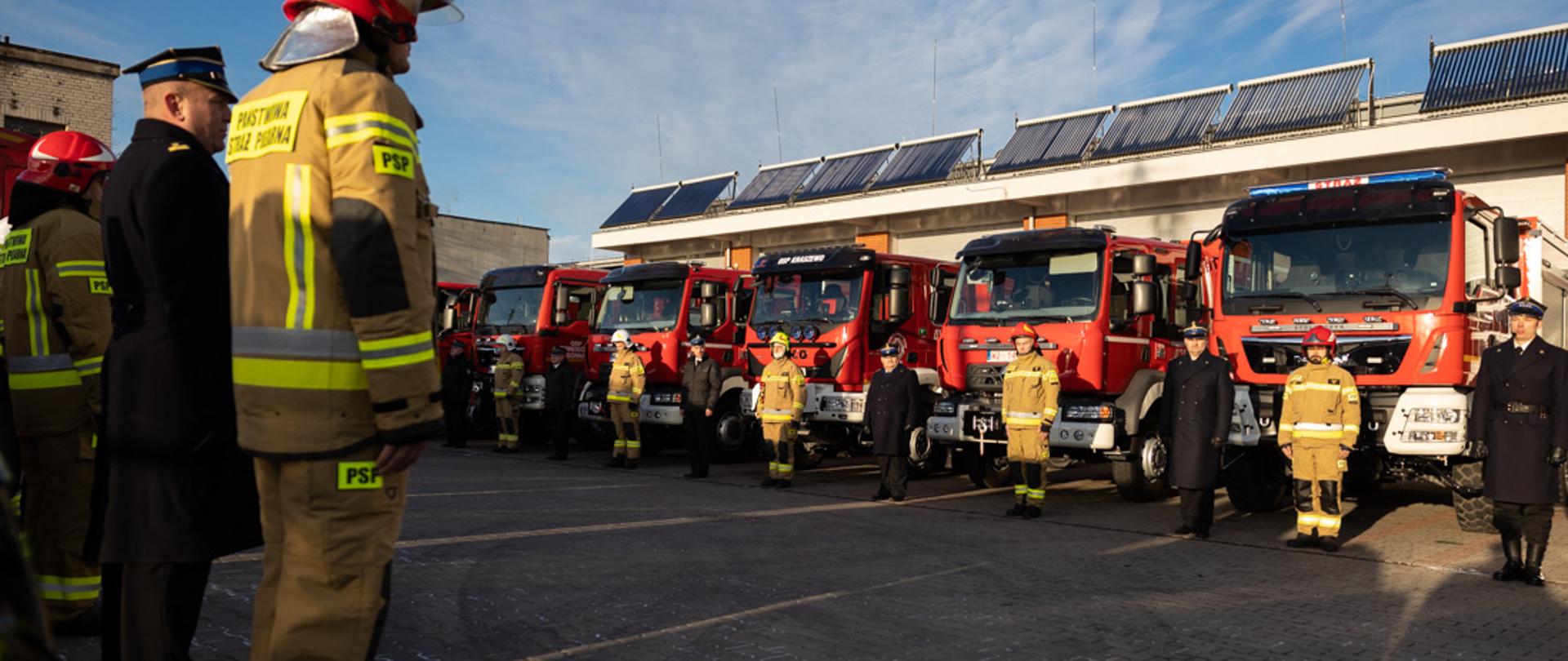 Widok na plac, na którym odbyła się uroczystość. Widoczne nowe samochody ratowniczo-gaśnicze oraz stojący przy nich strażacy.