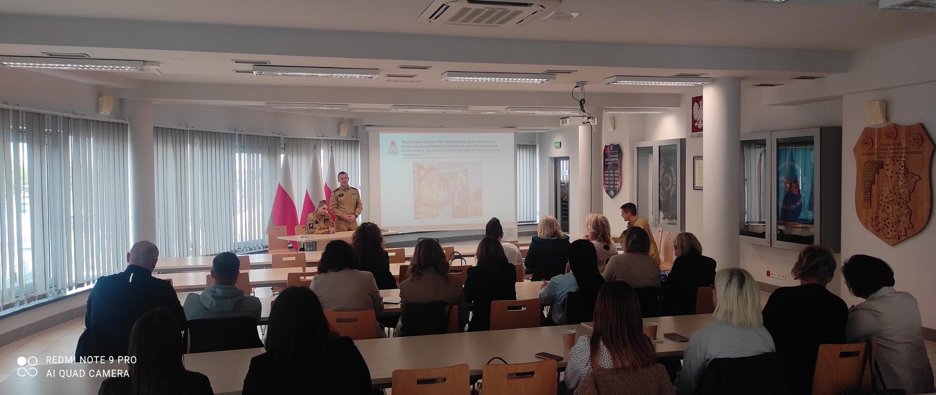 Sala konferencyjna wypełniona uczestnikami siedzącymi przy stołach, tyłem do obiektywu. Na środku przed ekranem projekcyjnym stoi dwoje strażaków w piaskowych mundurach służbowych, prowadzących prelekcję na tle flag Polski.