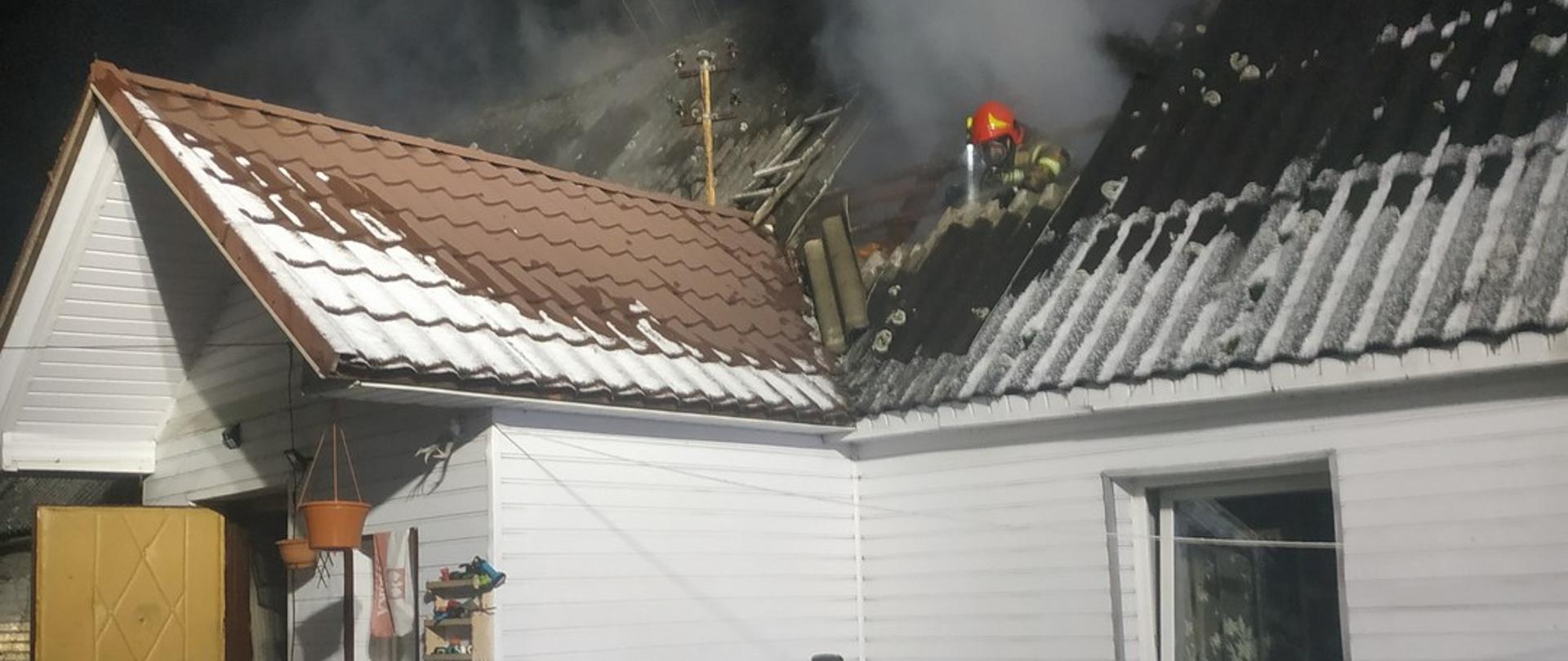 Strażak stoi na poddaszu budynku mieszkalnego, jest ubrany w czerwony hełm, ubranie specjalne koloru żółtego oraz założony ma na plecach aparat dróg oddechowych - prowadzi działania ratownicze.