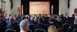 Widok ogólny z tyłu – zdjęcie zbiorowe. Uczestnicy uroczystości siedzą na sali GOK. Na drugim planie przodem po prawej stronie na scenie przemawia zastępca małopolskiego komendanta wojewódzkiego PSP. W tle wyświetlona plansza z nazwą wydarzenia.
