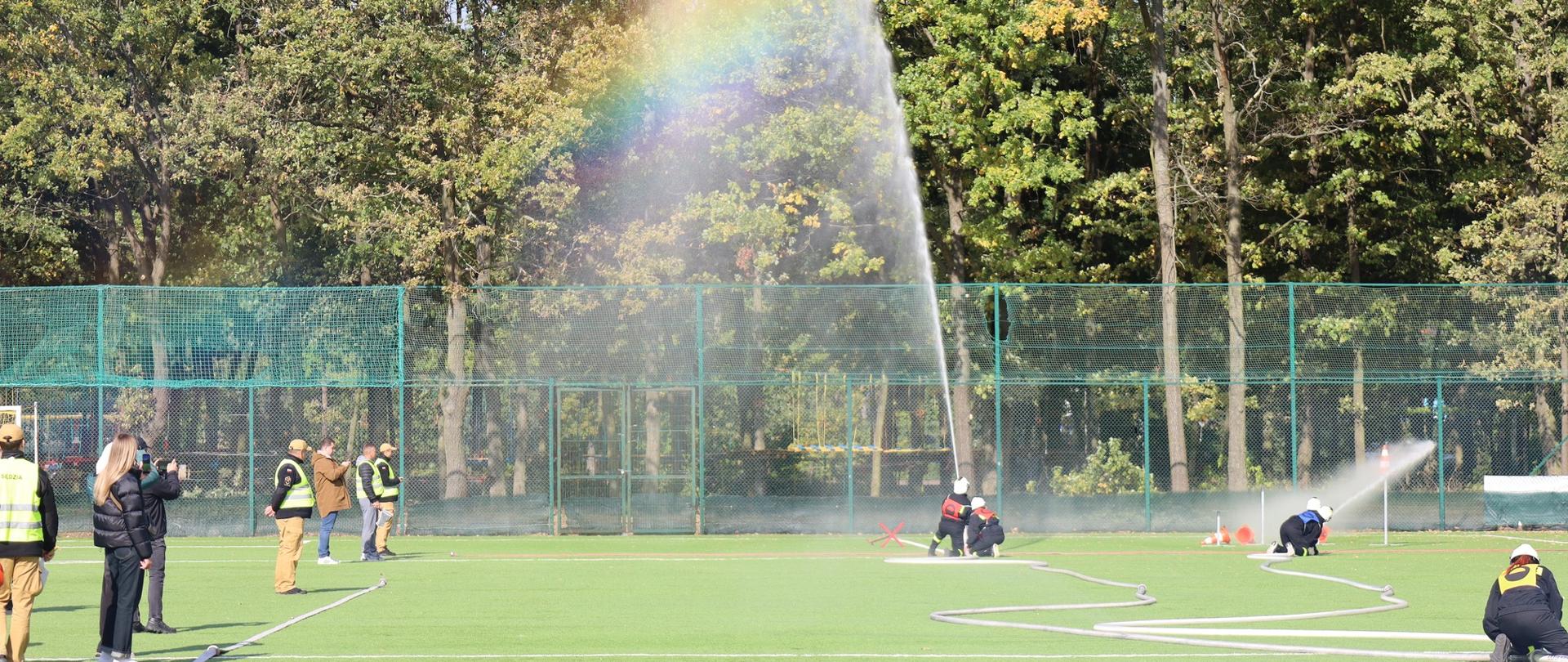 Na zdjęciu widać boisko sportowe, z zieloną nawierzchnią. Na środku kadru znajduje się dwóch zawodników strażaków, którzy trzymają prądownicę wodną, z której leci woda tworząc łuk, oraz tęczę. Po lewej stronie stoją ludzie, niektórzy w kamizelkach odblaskowych pełniących rolę sędziego oraz inni obserwujący zdarzenie. Po prawej stronie, widoczni są strażacy, którzy również biorą udział w zawodach, kierują oni strumieniem wody i obsługują węże pożarnicze. W tle widać drzewa i siatkę ogrodzeniową. Całość sugeruje, że są to zawody związane ze Sportem Pożarniczym.