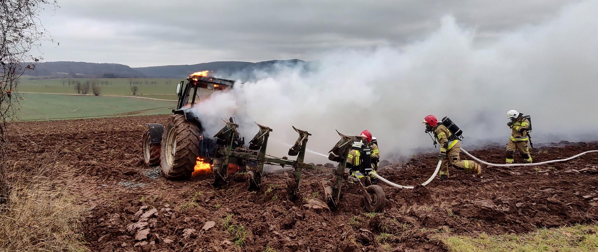 palący się ciągnika rolniczy na polu, za nim trzech strażaków gaszących pożar przy użyciu linii gaśniczej
