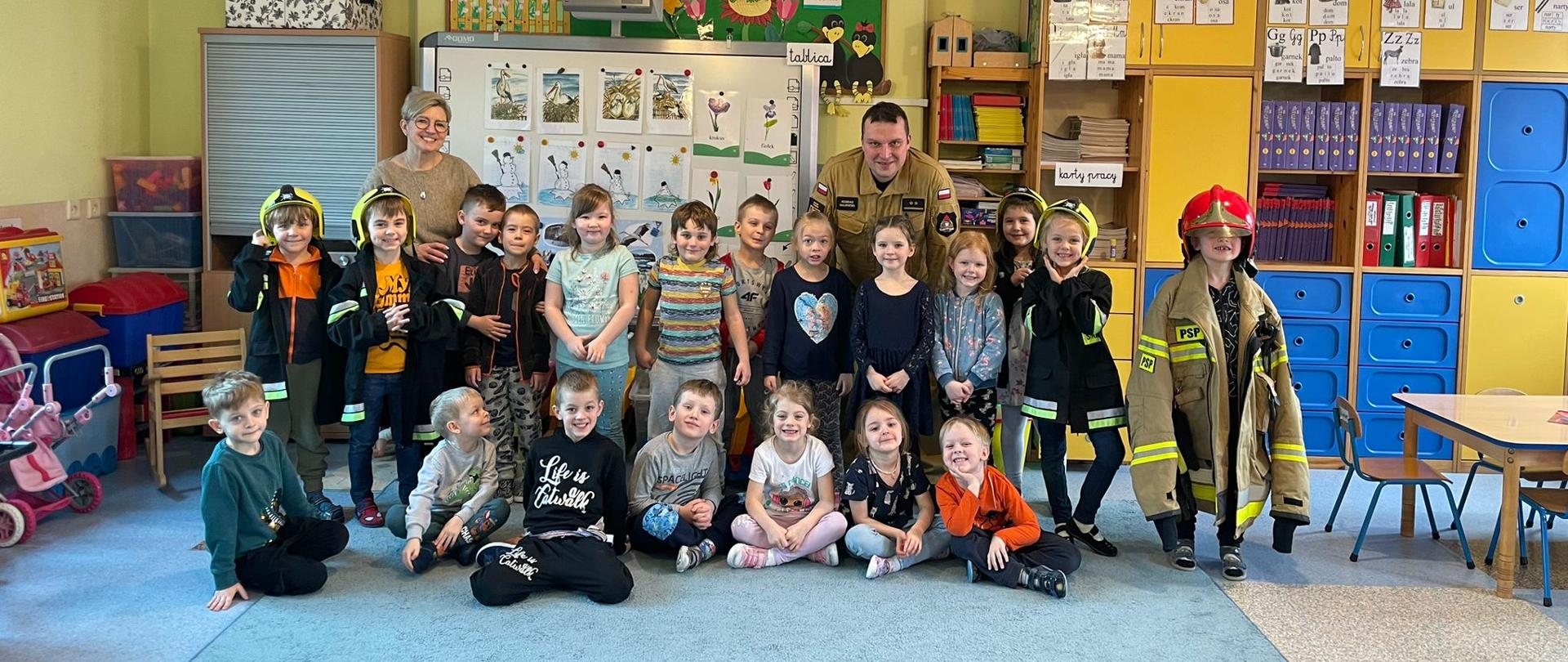 Zdjęcie grupowe w sali przedszkolnej z dziećmi w wieku około 6 lat - 20 dzieci. Na zdjęciu również Pani opiekunka i strażak PSP.