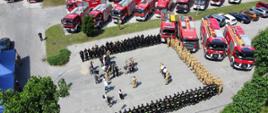 Widok z góry na plac na którym stoją w kształcie litery U ustawieni strażacy oraz z boku samochody PSP