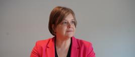 Wiceminister Zioło-Pużuk w czerwonej marynarce siedzi za stołem, za nią szara ściana.