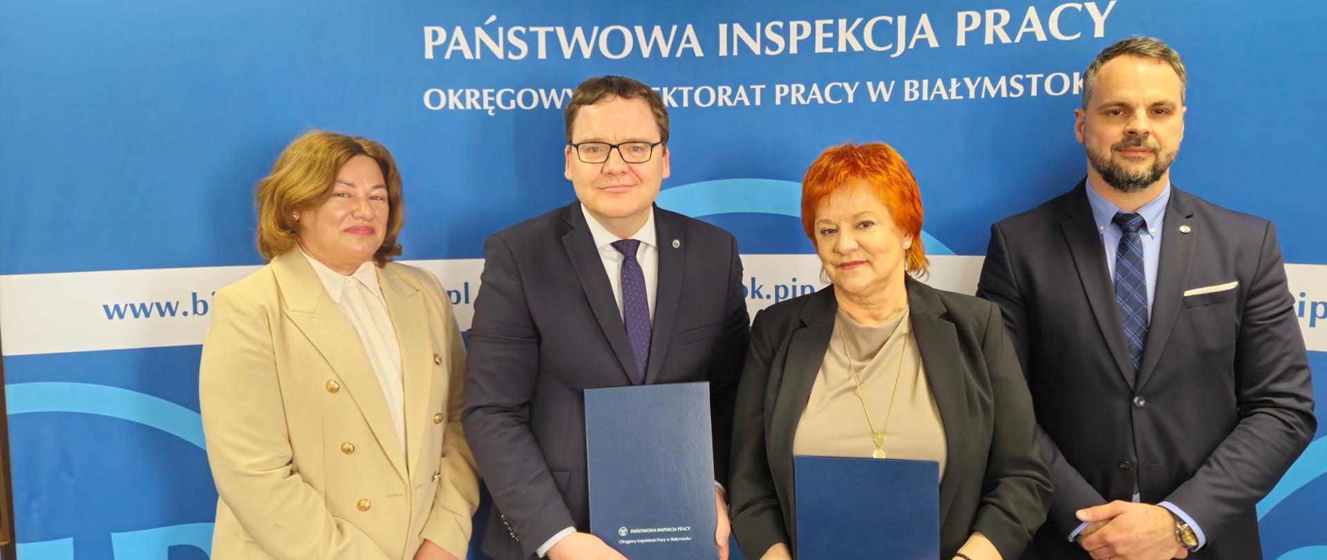4 osoby na tle banneru w kolorze niebieskim z logo i napisem Państwowa Inspekcja Pracy. Od lewej strony: Pani Małgorzata Kruszewska Kierownik Oddziału Higieny Pracy i Nadzoru Nad Chemikaliami w Wojewódzkiej Stacji Sanitarno-Epidemiologicznej w Białymstoku, Pan dr Karol Łapiński p.o. Okręgowego Inspektora Pracy w Białymstoku, Pani dr n. med. Maria Siewko – Podlaski Państwowy Wojewódzki Inspektor Sanitarny w Białymstoku, Pan Tomasz Werdoni p.o. Zastępcy Okręgowego Inspektora Pracy ds. Nadzoru w Białymstoku