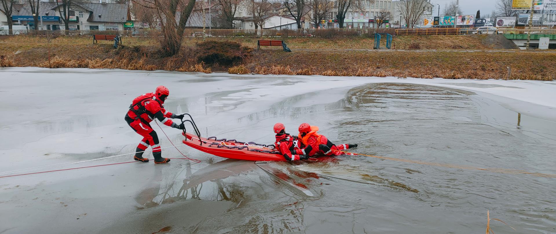 Zamarźnięty staw z przeręblem. Dwóch strażaków ubranych w skafandry ratownicze przy pomocy czerwonych sań lodowych ćwiczą wydobywanie osoby pod którą załamał się lód.