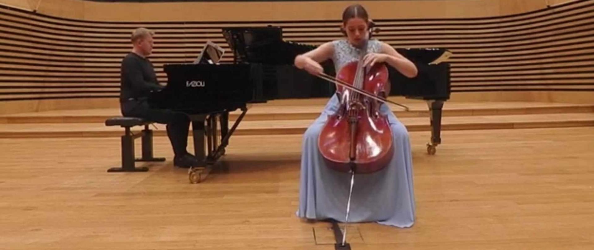Nastolatka gra na wiolonczeli na środku estrady sali koncertowej PSM, za nią na fortepianie gra mężczyzna.