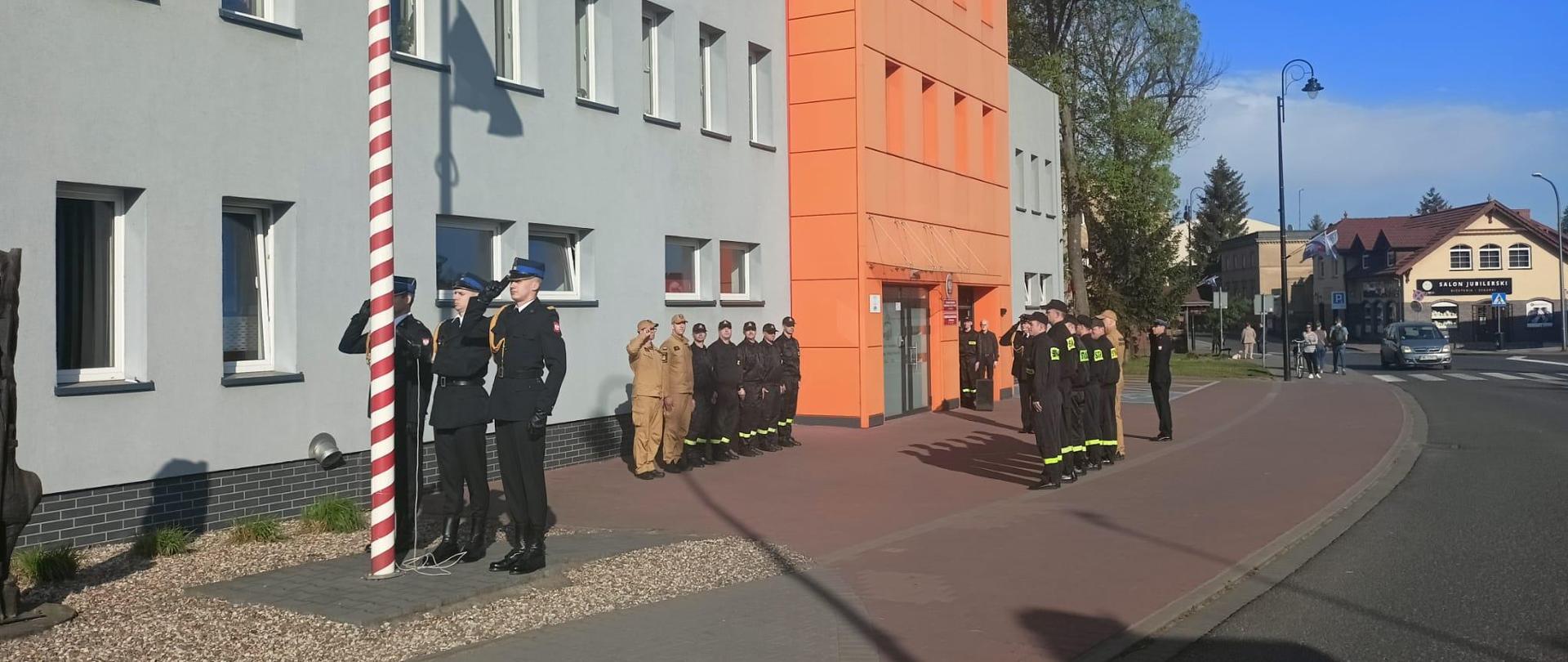 Zdjęcie wykonane na zewnątrz. Słoneczna pogoda. Na zdjęciu widoczni strażacy podczas uroczystej zbiórki z okazji Dnia Flagi państwowej RP. W tle budynek Komendy.