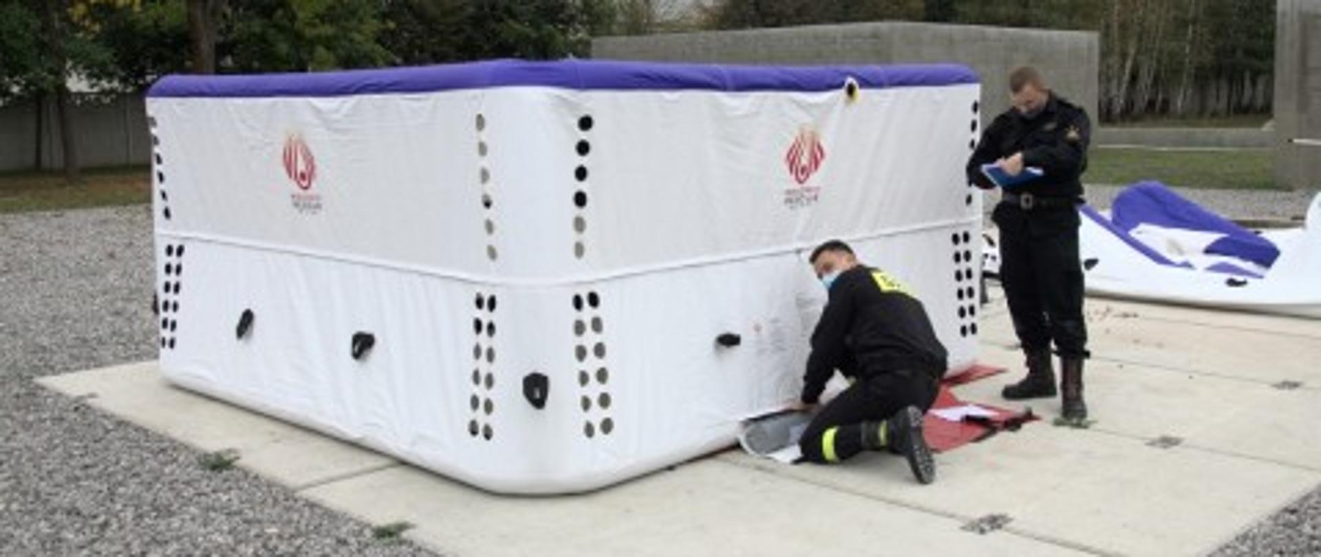 Na zewnątrz na betonowej płycie znajduje się napompowany skokochron. Obsługuje go jeden strażak, natomiast drugi stoi obok i notuje coś w zeszycie. W oddali widać drzewa/