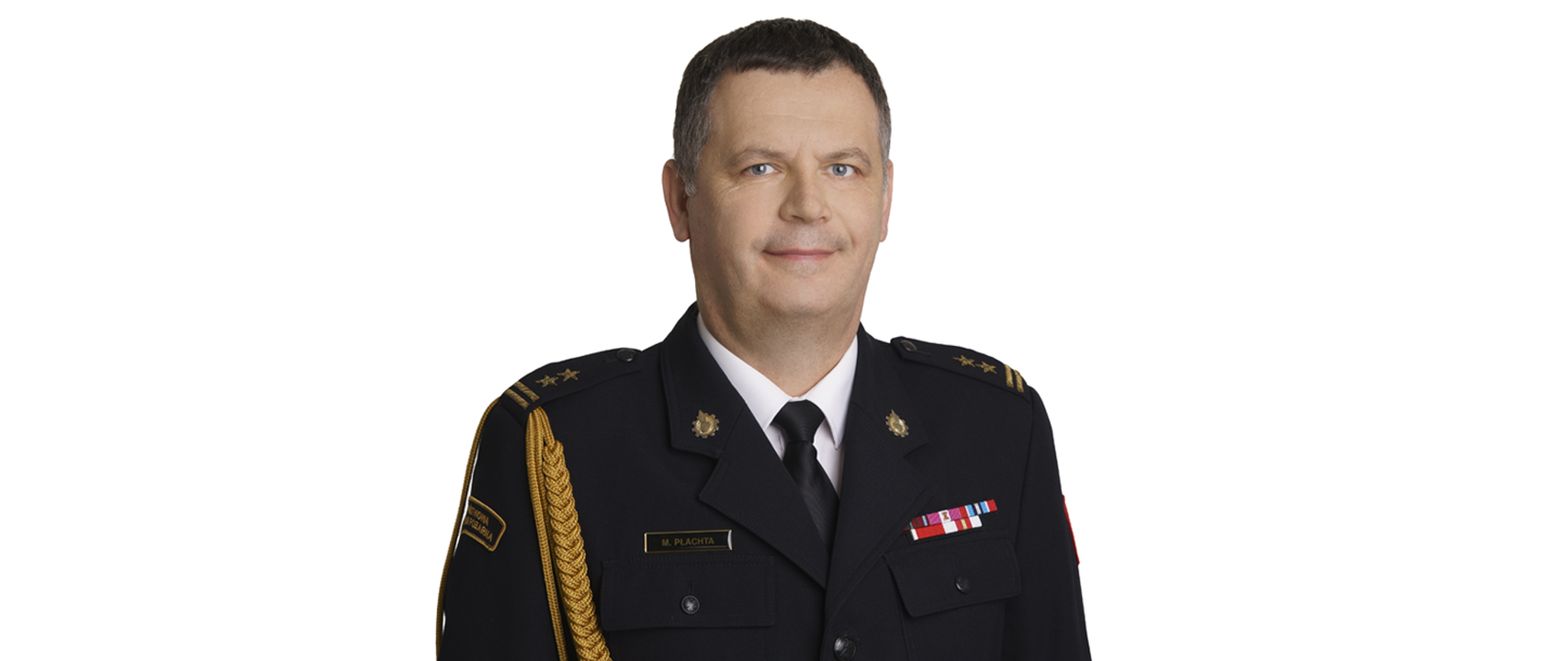 Na zdjęciu widoczny jest portret Marcina Płachty, zastępcy komendanta, ubranego w granatowy mundur galowy Państwowej Straży Pożarnej. Ma krótkie, ciemne włosy i patrzy prosto w obiektyw, uśmiechając się lekko. Na ramionach munduru ma złote sznury, a na pagonach widoczne są gwiazdki. Na piersi ma przypięte odznaczenia i plakietkę z nazwiskiem.