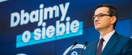 Premier Mateusz Morawiecki na tle ścianki wizyjnej.