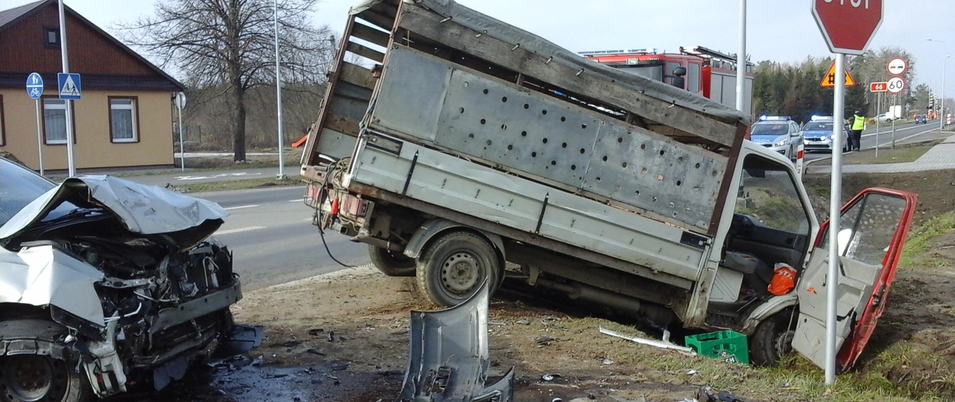 Samochód dostawczy stojący na poboczu po zderzeniu z innym pojazdem. Przód samochodu znajduje się w przydrożnym rowie. Przed nim widoczny rozbity przód drugiego pojazdu biorącego udział w zdarzeniu. W tle wóz strażacki oraz dwa radiowozy