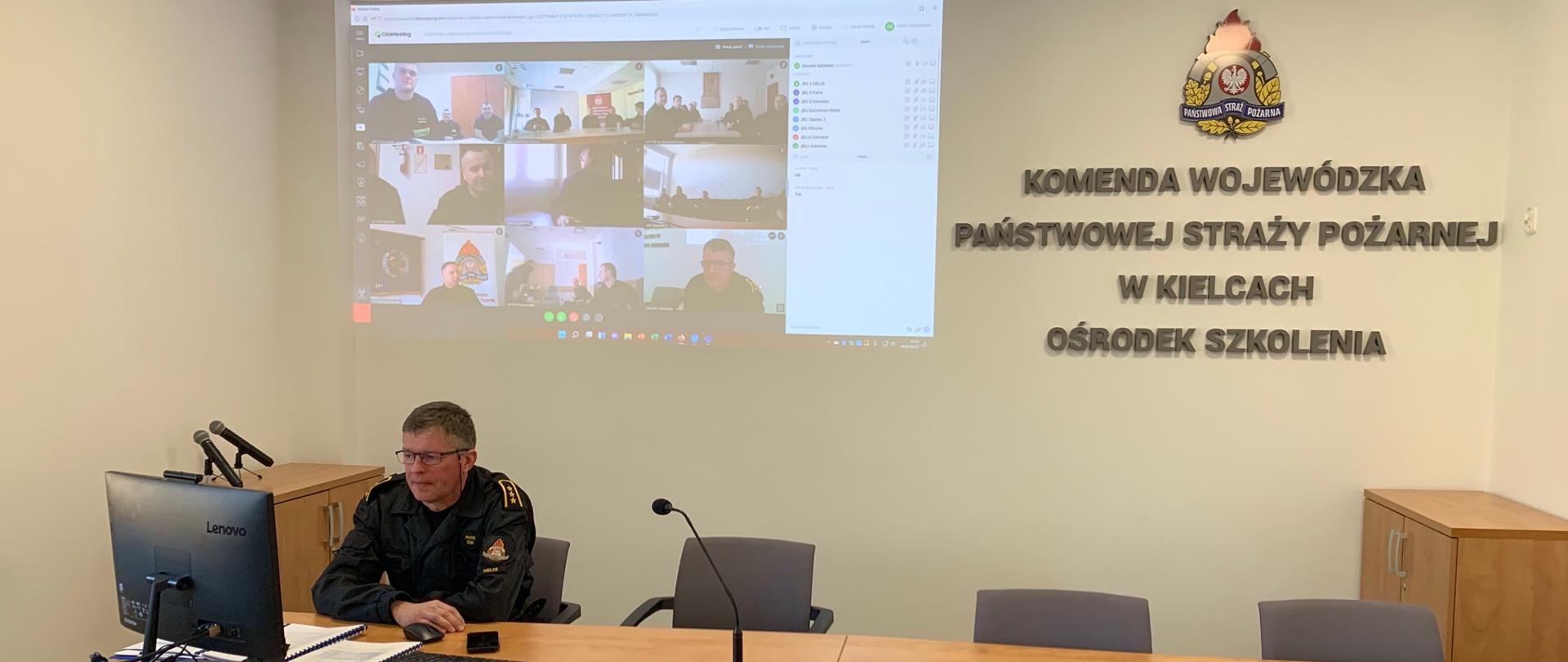Zdjęcie zrobione jest w sali narad. Na pierwszym planie zdjęcia widać siedzącego na krześle, ubranego w czarną odzież mężczyznę prowadzącego zajęcia. Mężczyzna skierowany jest w naszym kierunku.
Patrzy w monitor stojący przed nim. Na drewnianym biurku stoi mikrofon, akcesoria komputerowe oraz notatki do zajęć.
Za nim na białej ścianie widnieje napis oraz logo Państwowej Straży Pożarnej.
Obok napisu wyświetlany jest obraz rzucany projektorem a na nim postacie osób uczestniczących w szkoleniu.
