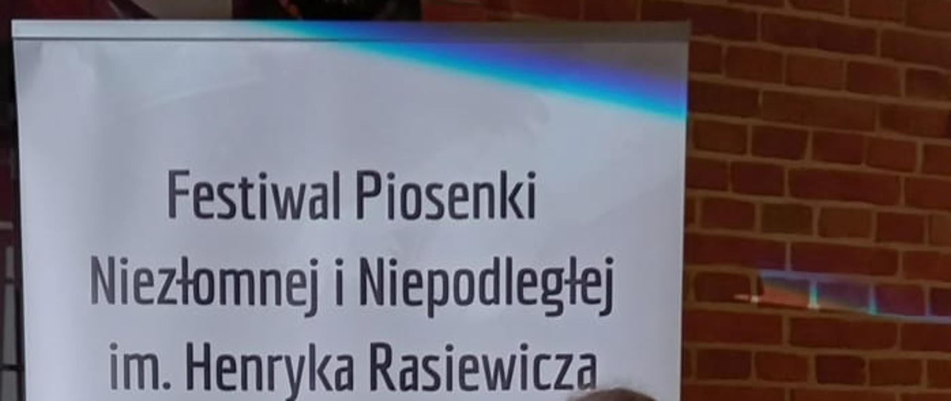 Zdjęcie z VII Ogólnopolskiego Festiwalu Piosenki Niezłomnej i Niepodległej w Krakowie. Na zdjęciu uczennica stoi przy biało-czerwonym rollupie z nazwa konkursu. Dziewczynka ubrana jest w czarne spodnie i buty oraz białą bluzkę, we włosach ma czerwoną wstążkę, w rękach trzyma dyplom i statuetkę. W tle ściana pokryta cegłą.