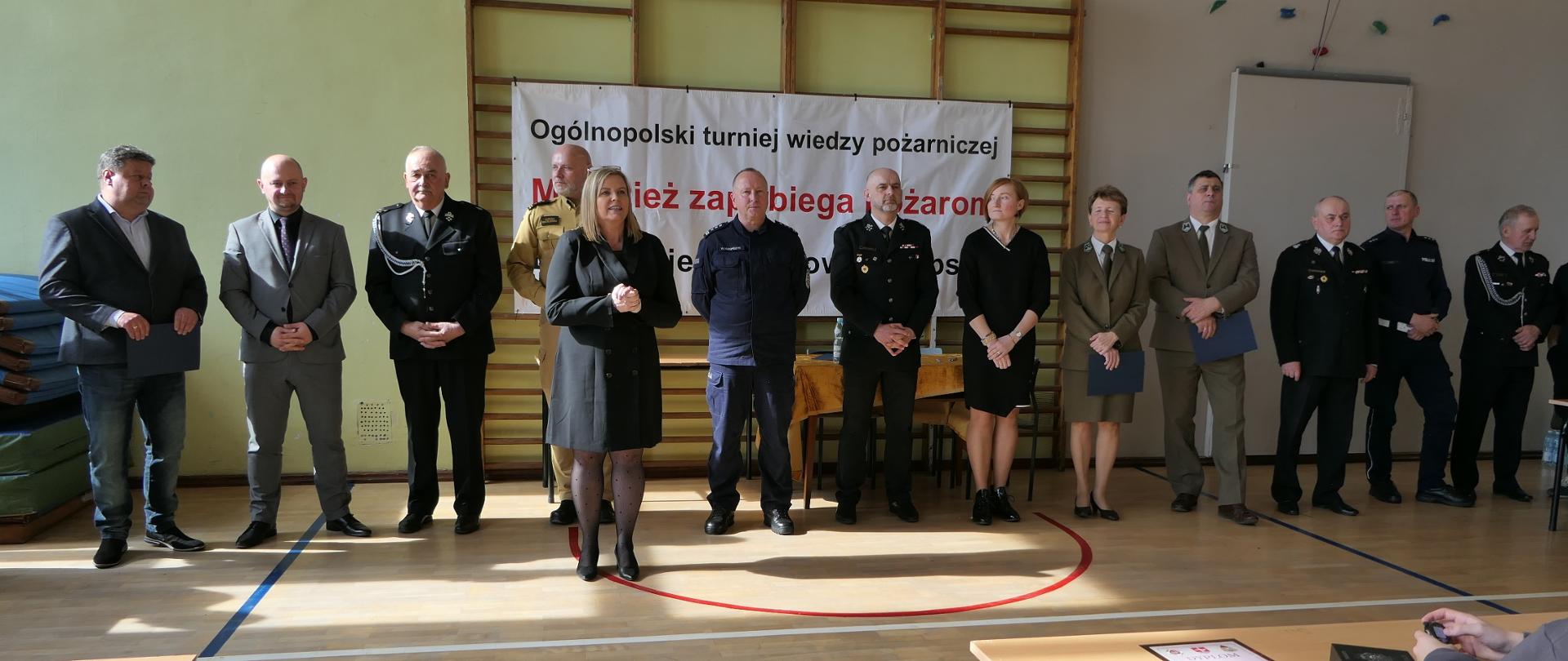 Turniej OTWP 2025 zaproszeni goście oraz organizatorzy. Zdjęcie przedstawia grupę dorosłych osób stojących w rzędzie na tle białego baneru z napisem "Ogólnopolski turniej wiedzy pożarniczej". Na banerze widnieje również fragment tekstu "Młodzież zapobiega pożarom".
Wszyscy stojący na pierwszym planie są elegancko ubrani. Niektórzy z nich mają na sobie garnitury, inni mundury strażackie. W centrum grupy, na pierwszym planie, stoi kobieta w czarnym płaszczu. Tło stanowi ściana sali gimnastycznej z panelami, a na podłodze widoczne są oznaczenia boiska.