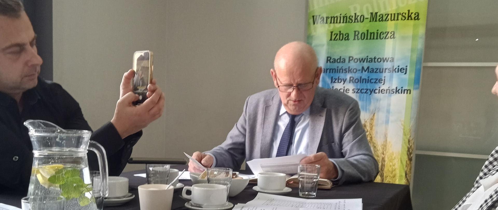 Mężczyzna – przewodniczący spotkania, siedzi przy stole, za sobą ma baner na stojaku z napisem
Warmińsko-Mazurska Izba Rolnicza