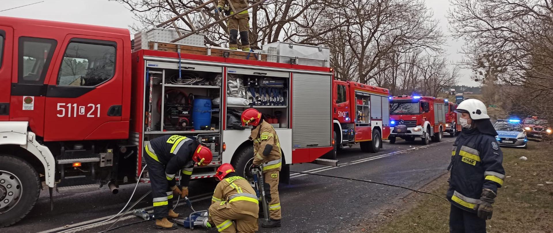Cztery samochody pożarnicze stoją na jezdni. Strażacy przygotowują sprzęt hydrauliczny do działań. W tle po prawo widoczny radiowóz a za nim jeszcze jeden osobowy pojazd pożarniczy.