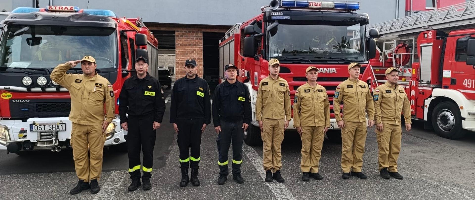 Na zdjęciu znajdują się strażacy ustawieni do zdjęcia na tle samochodów bojowych oraz budynku komendy
