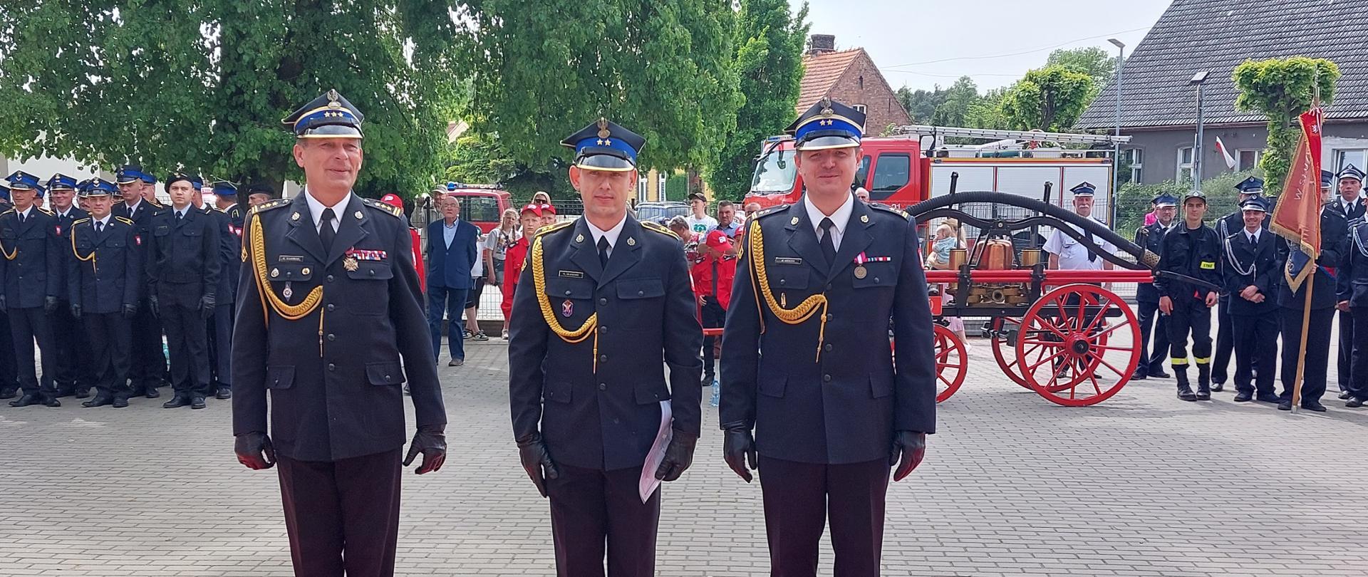 trzech strażaków stoi na baczność na czerwonym dywanie, w tle pompa pożarnicza oraz grupa strażąków 